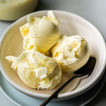 A white bowl contains three scoops of low carb, protein ice cream, accompanied by a metal spoon. A jar with a small amount of a white substance is visible in the background. Made with the Ninja Creami, this dessert is both delicious and nutritious.
