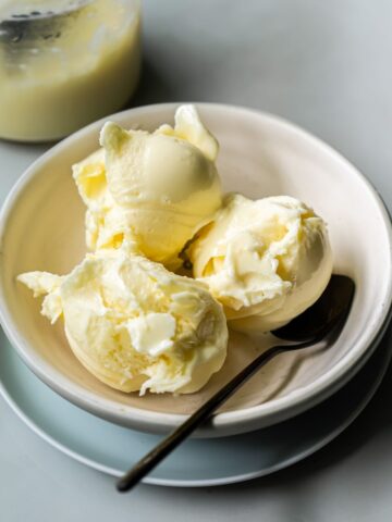 A white bowl contains three scoops of low carb, protein ice cream, accompanied by a metal spoon. A jar with a small amount of a white substance is visible in the background. Made with the Ninja Creami, this dessert is both delicious and nutritious.