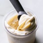 Close-up of creamy white Ninja Creami Oat Milk Coconut Protein Ice Cream in a clear pint, with a black spoon lifting a smooth swirl, showcasing its rich texture and consistency.