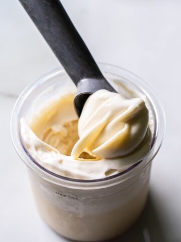 Close-up of creamy white Ninja Creami Oat Milk Coconut Protein Ice Cream in a clear pint, with a black spoon lifting a smooth swirl, showcasing its rich texture and consistency.