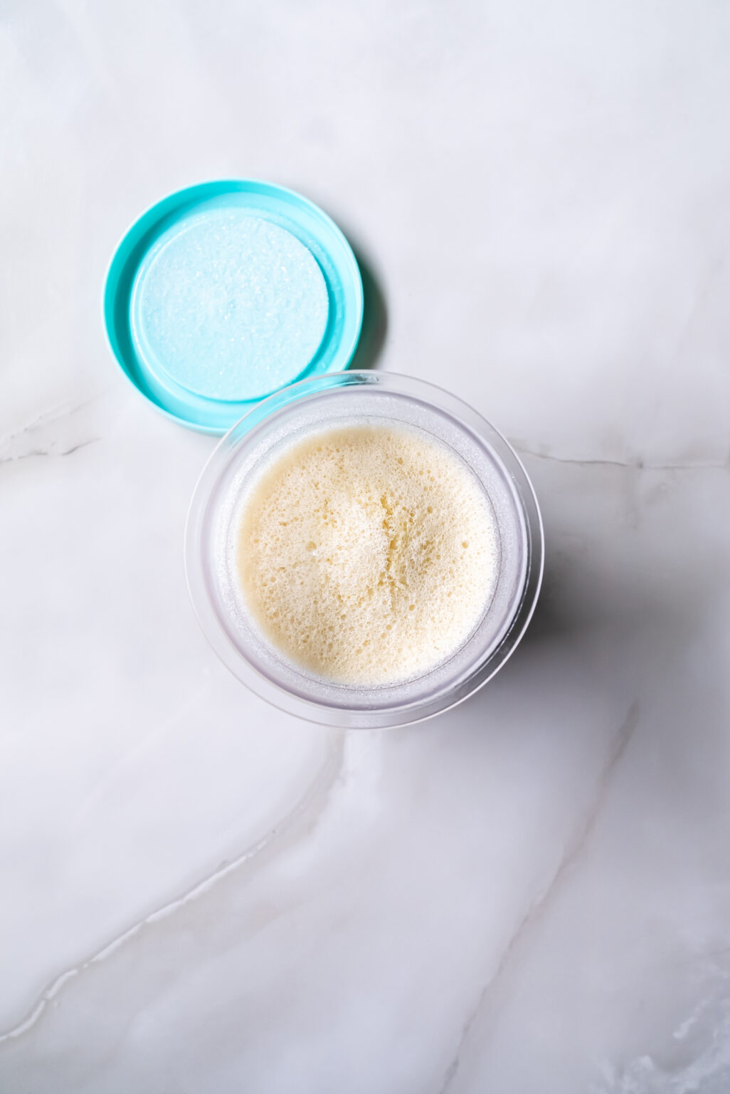 A close-up view of a Ninja Creami pint filled with frozen frothy liquid, perfect for whipping up ice cream, with its lid placed beside it on a white surface.