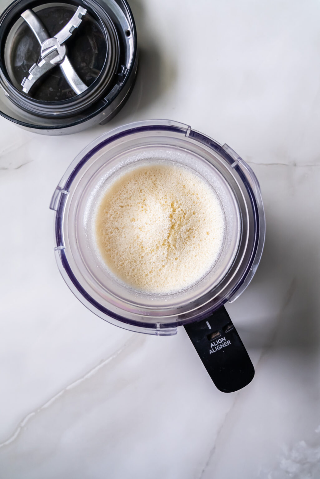 A clear plastic food processor bowl filled with frozen Coconut protein mix sits on a white countertop, ready to become the base for delicious ice cream using Ninja Creami.