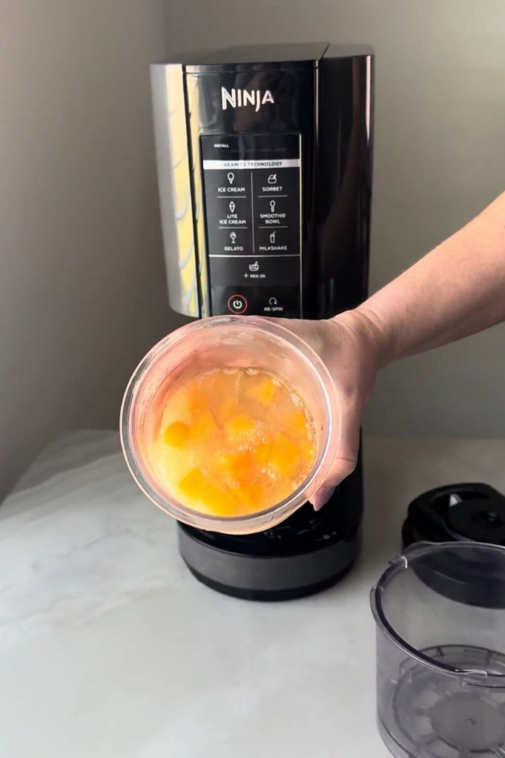 Hand holding a frozen Ninja Creami pint containing solidified peach mixture above the machine’s base, preparing to insert it for spinning into creamy Ninja Creami Peach Sorbet.