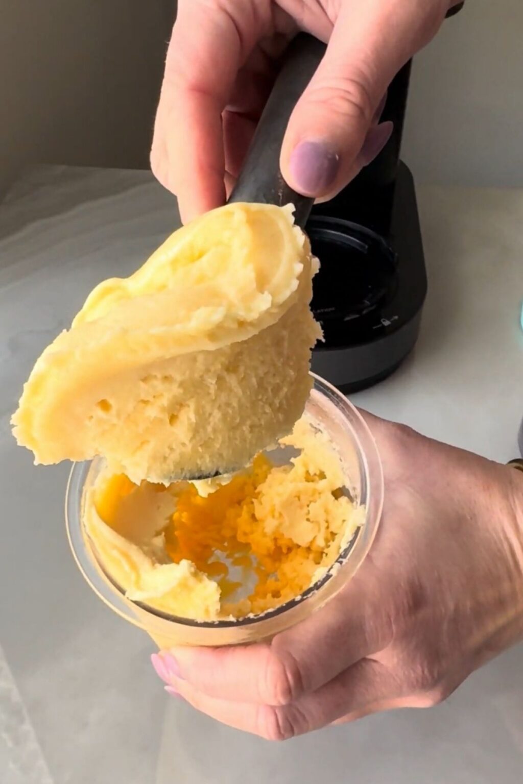 Hands using an ice cream scoop to lift a generous portion of bright yellow-orange Ninja Creami Peach Sorbet from the pint, showcasing its soft-serve-like texture and rich fruit flavor.