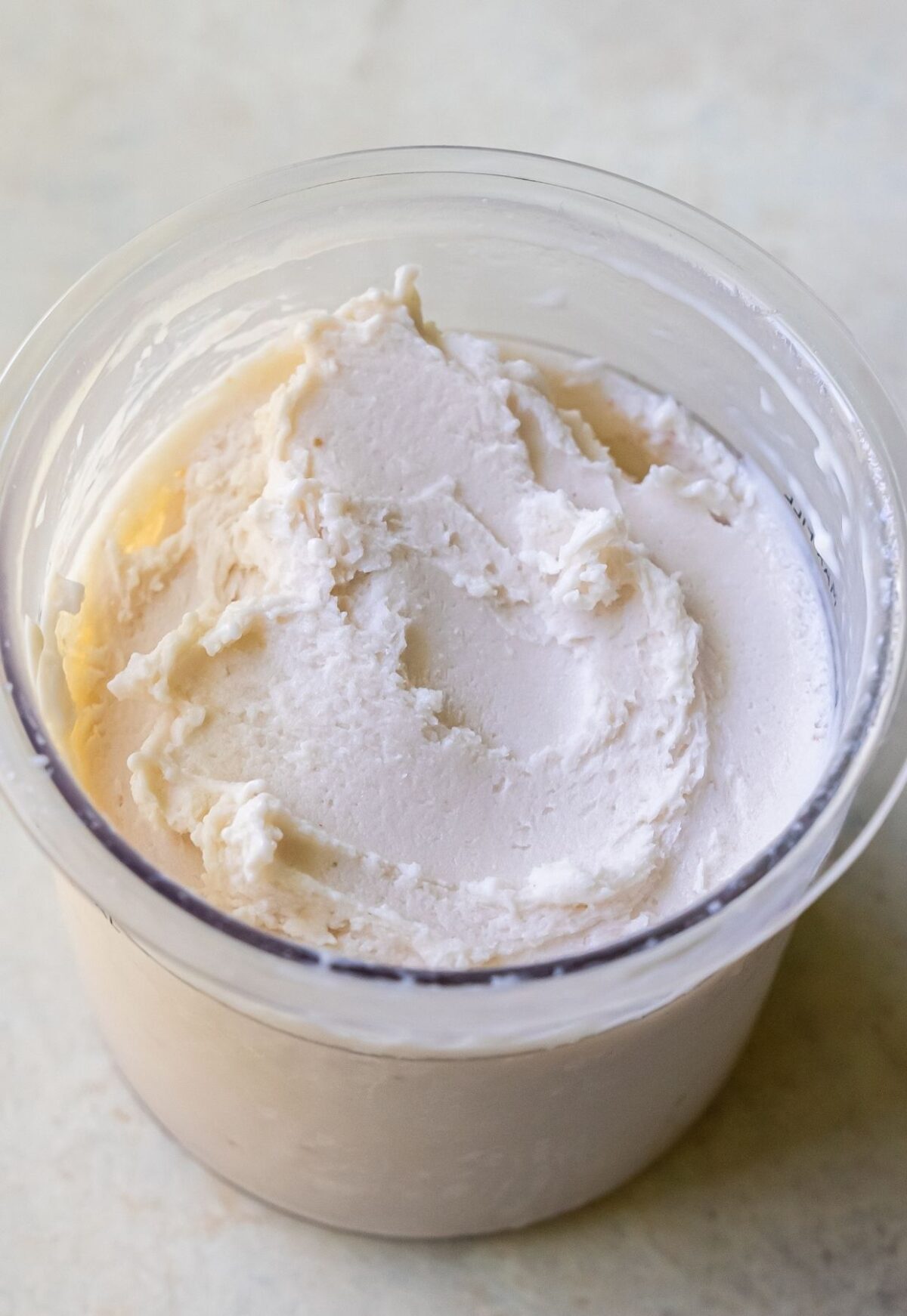 A container filled with a creamy, white soft substance, resembling frosting or whipped cream, sits against a plain background, evoking the rich texture of a Strawberry Cheesecake crafted in the Ninja Creami.