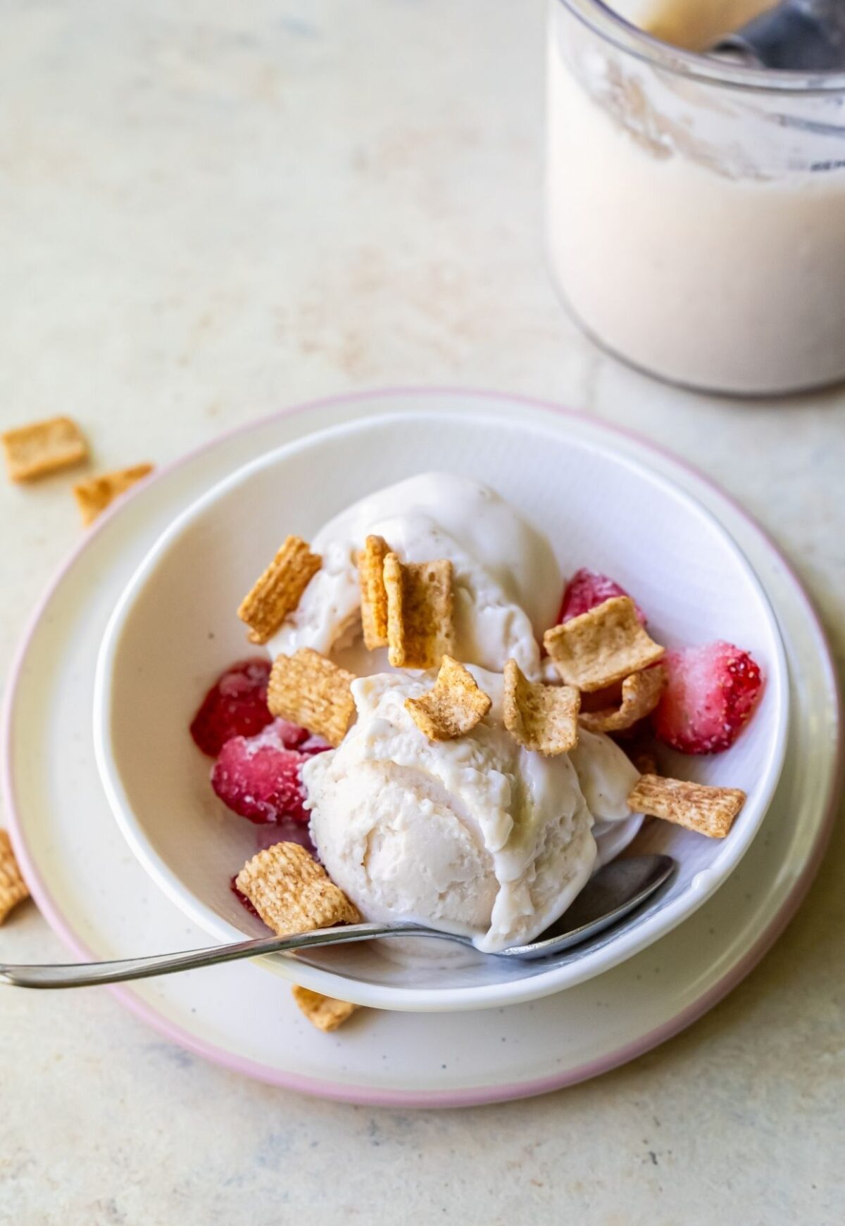 A bowl of protein ice cream topped with cereal squares and strawberry slices, with a spoon placed inside, is seen on a white plate. A glass container with more ice cream is in the background.