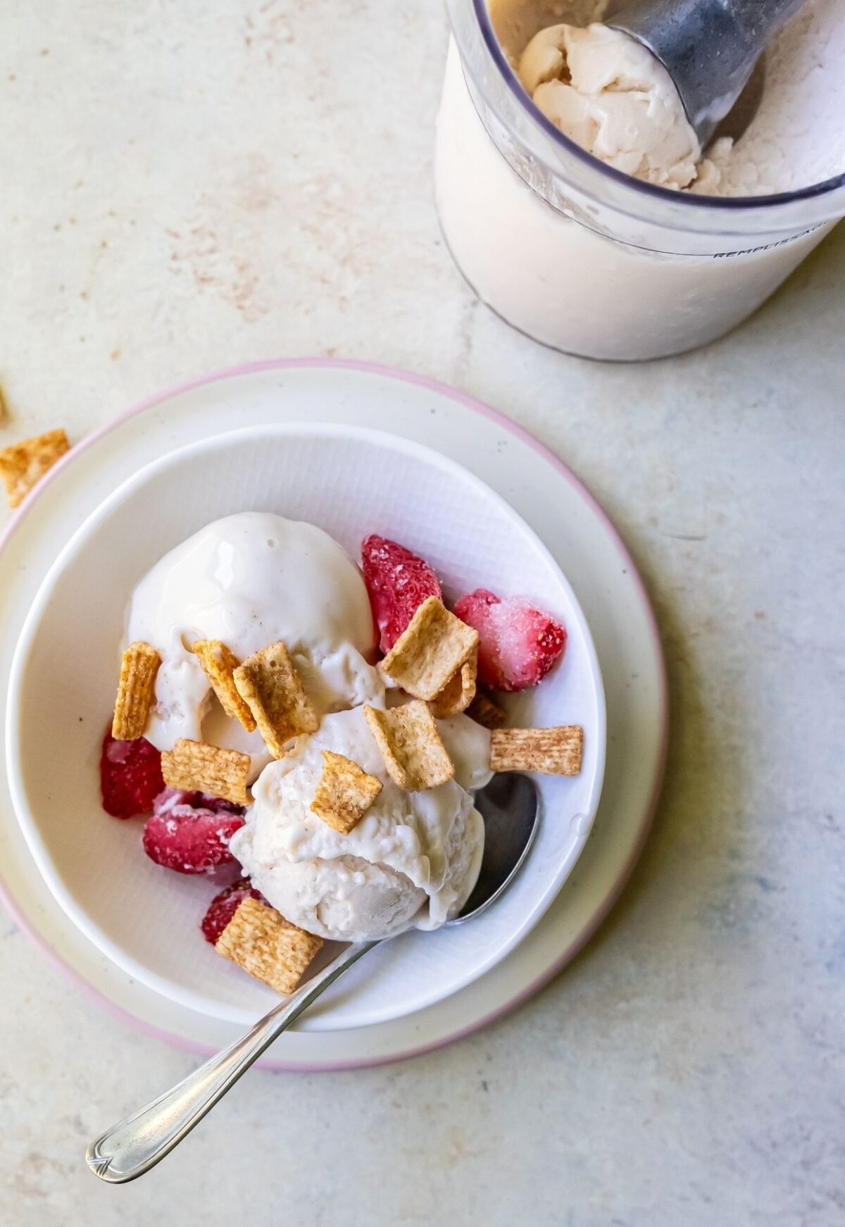 A bowl of vanilla protein ice cream topped with strawberries and cereal pieces, placed on a white plate with a spoon. A container with more ice cream, likely made with the Ninja Creami, is visible in the background.