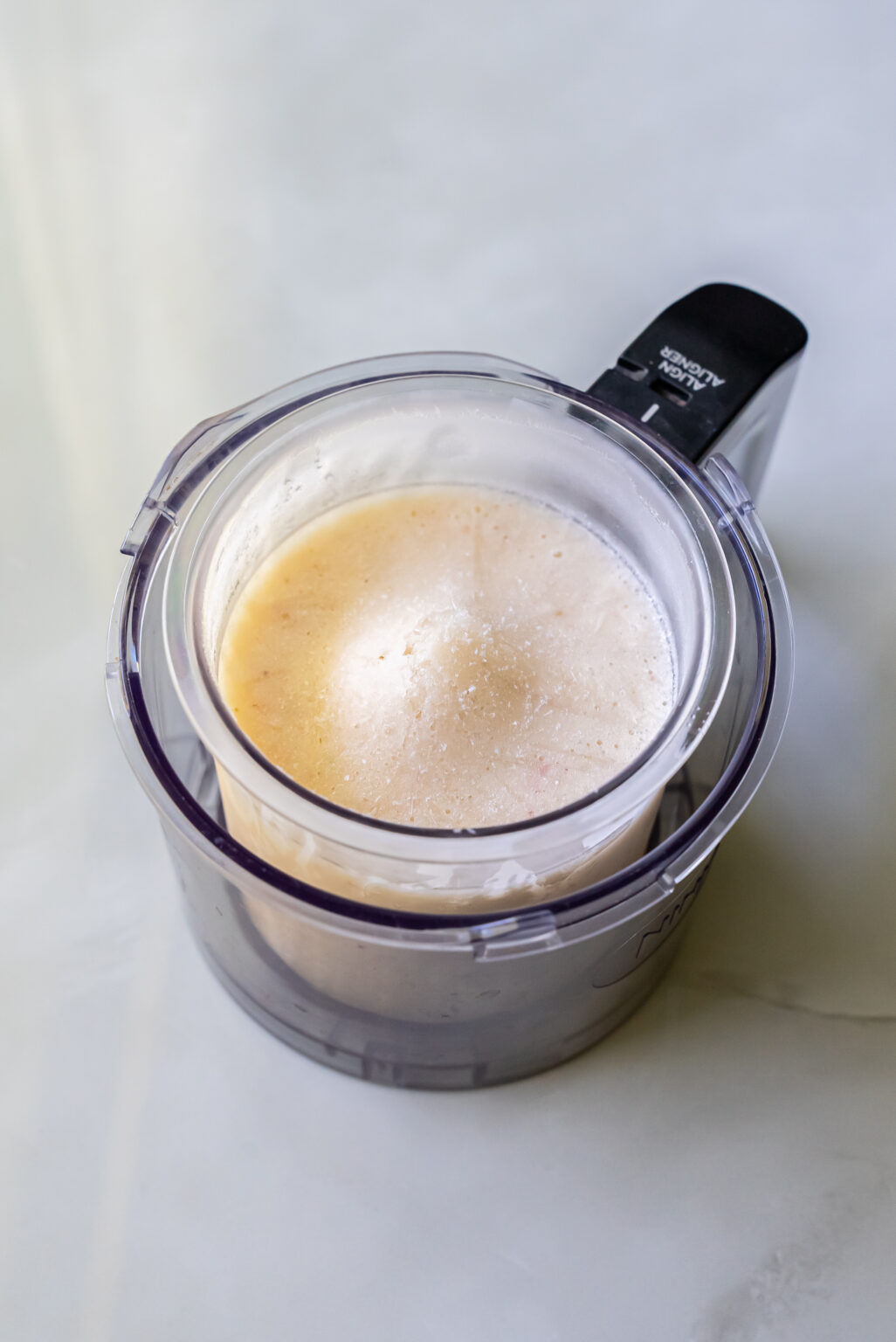 A food processor bowl filled with light brown creamy mixture, resembling the base for a decadent strawberry cheesecake, is placed on a light gray surface.