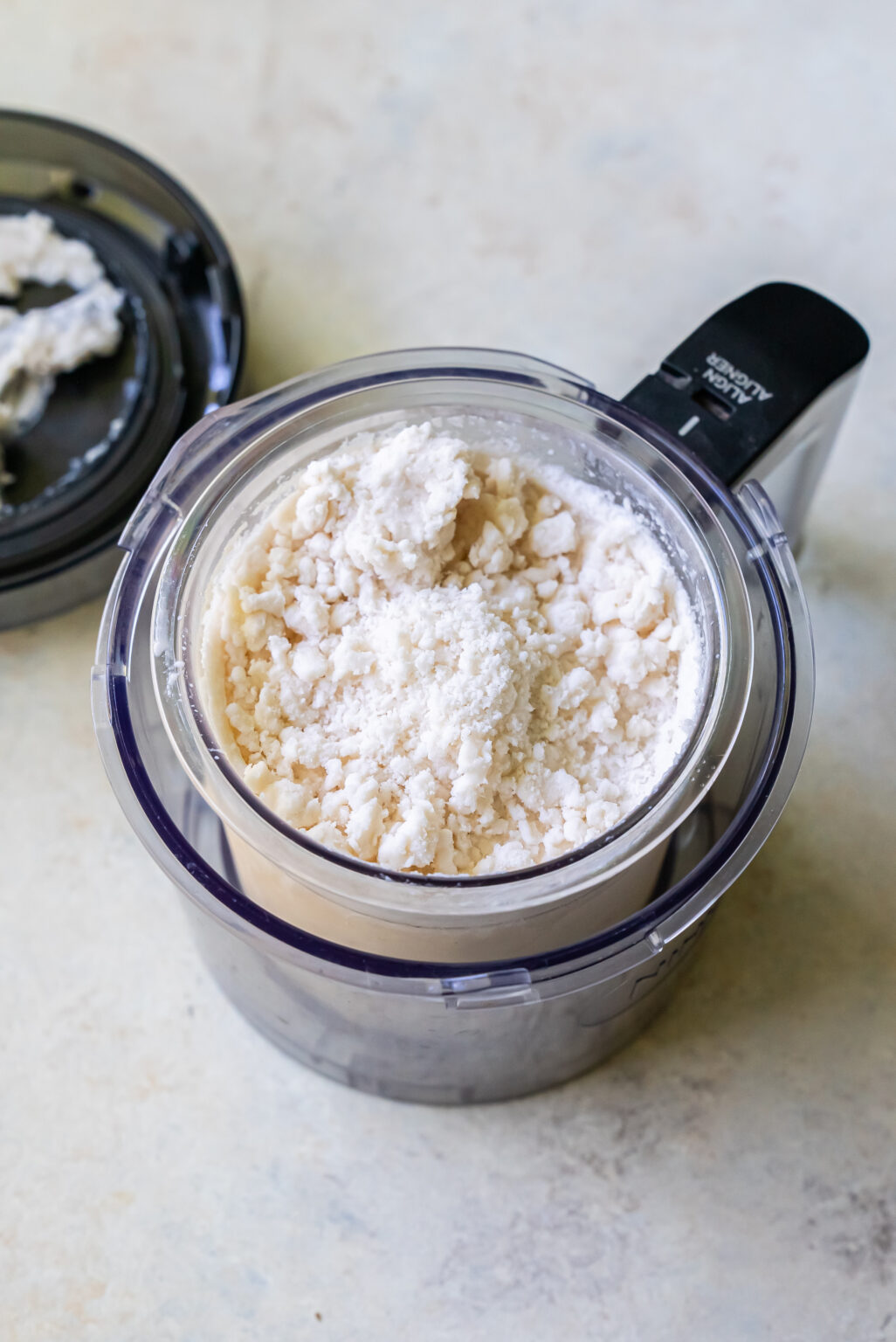 A clear food processor bowl filled with crumbled white cheese, ideal for creating a luscious Strawberry Cheesecake, with the lid removed and placed next to it.