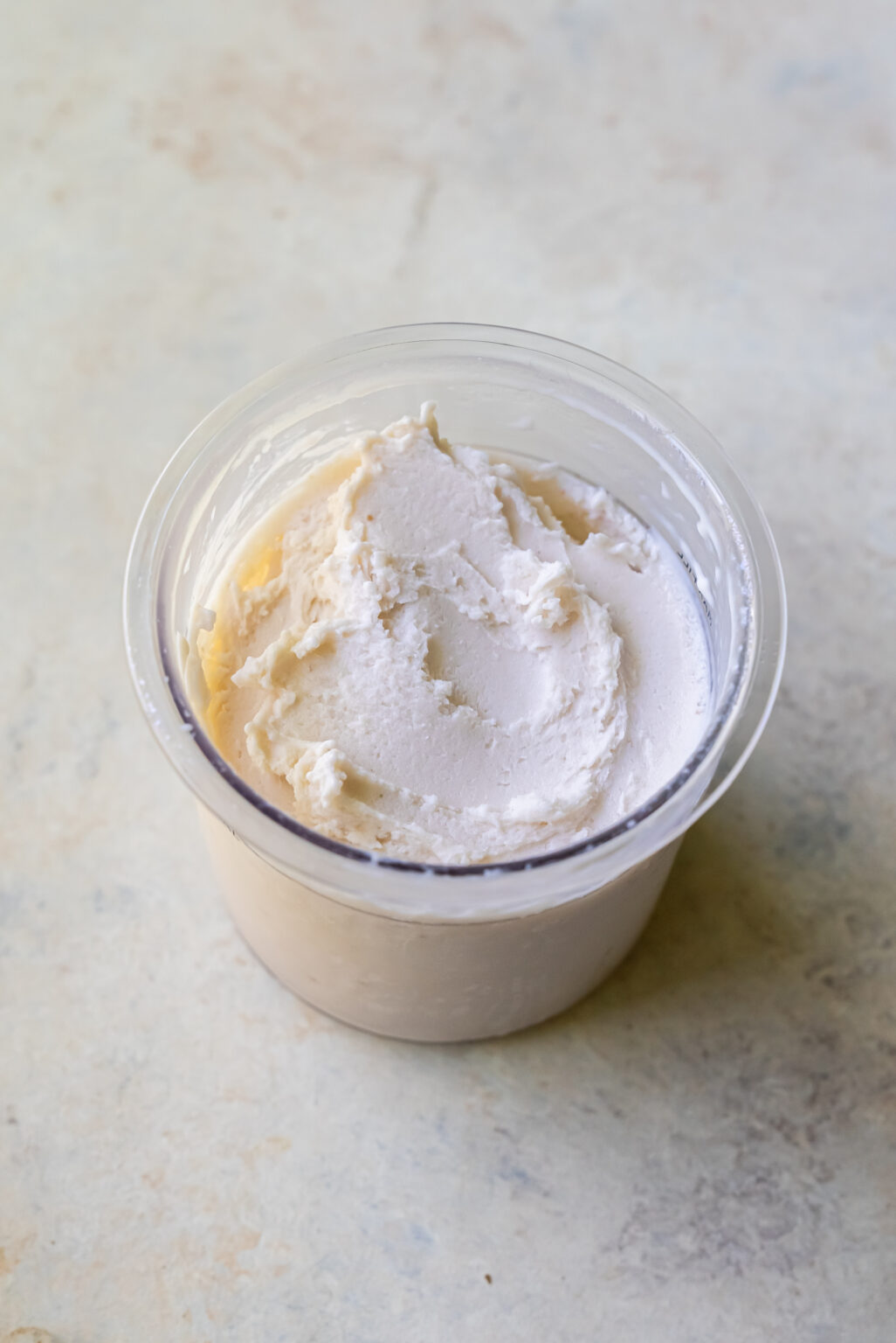 A clear plastic container filled with a smooth, white creamy substance, likely a type of dairy product, sits on a light-colored surface. This tempting treat might just be a delicious batch of Strawberry Cheesecake Protein Ice Cream made with the Ninja Creami.