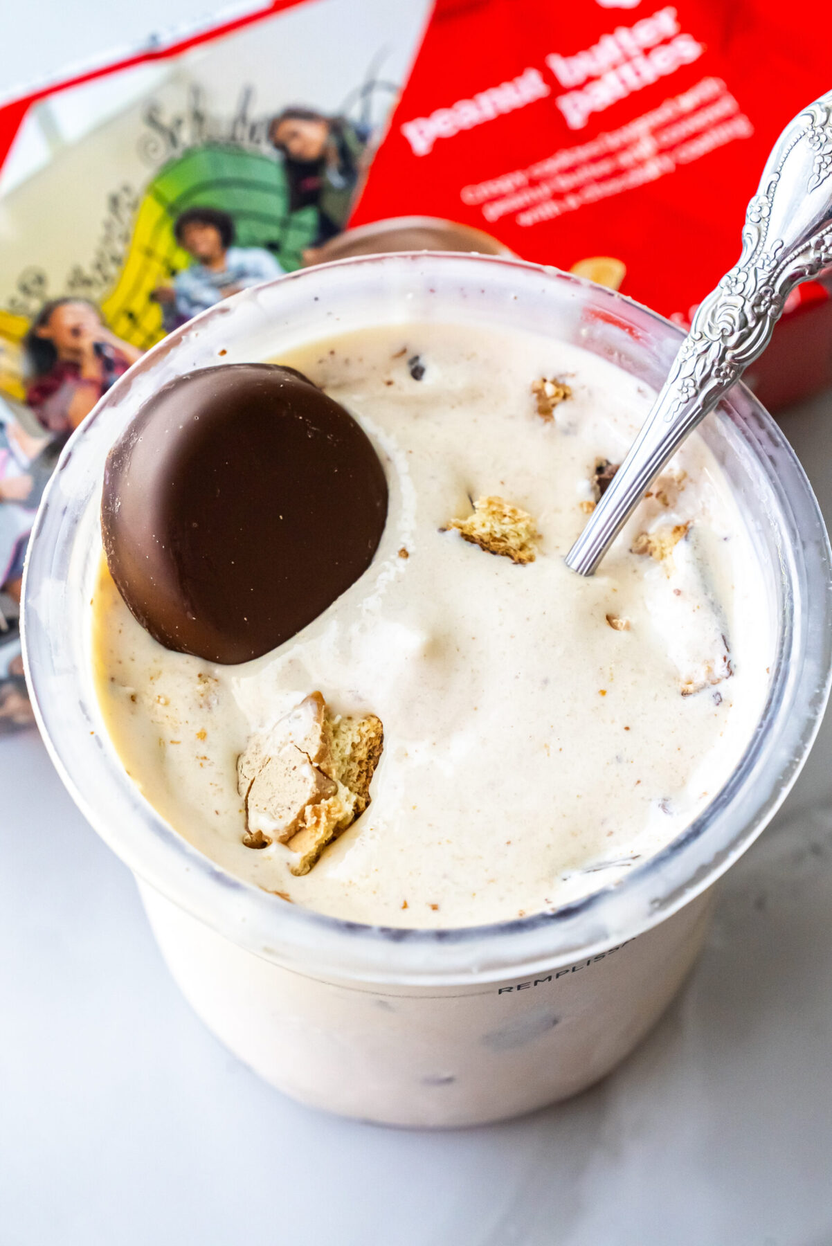 Top-down view of Peanut Butter Patties Ninja Creami Ice Cream in a clear pint. A whole chocolate-covered cookie sits on top alongside broken pieces, with an ornate silver spoon inserted into the mixture.