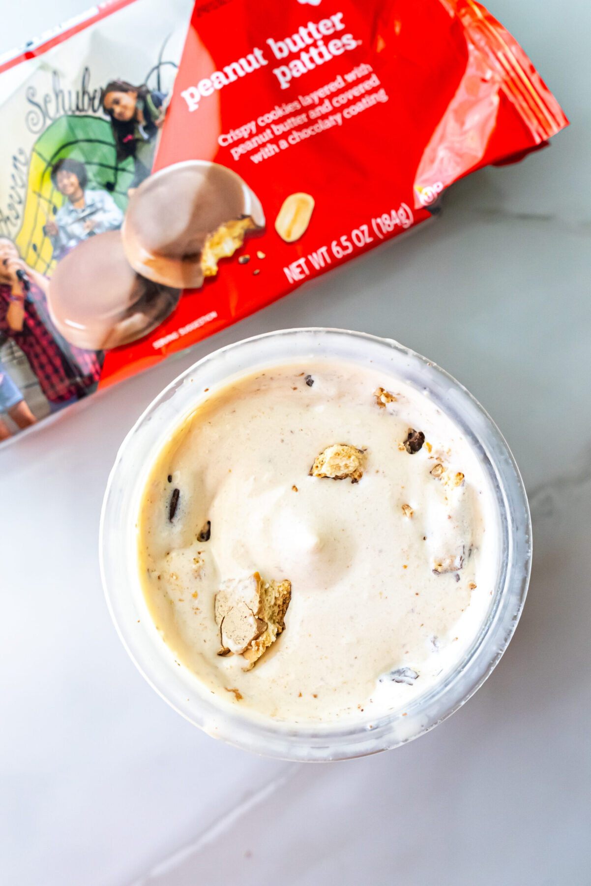 Overhead shot of Peanut Butter Patties Ninja Creami Ice Cream in a clear pint with broken cookie pieces mixed in. A red bag of Girl Scout Peanut Butter Patties lies diagonally behind the container on a white surface.