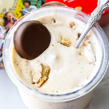 Top-down view of Peanut Butter Patties Ninja Creami Ice Cream in a clear pint. A whole chocolate-covered cookie sits on top alongside broken pieces, with an ornate silver spoon inserted into the mixture.