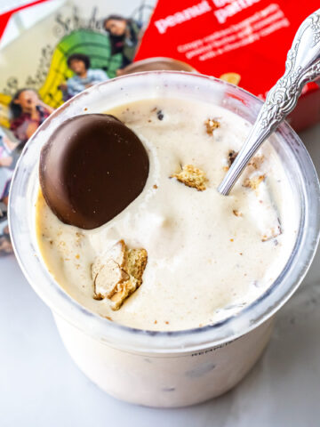 Top-down view of Peanut Butter Patties Ninja Creami Ice Cream in a clear pint. A whole chocolate-covered cookie sits on top alongside broken pieces, with an ornate silver spoon inserted into the mixture.