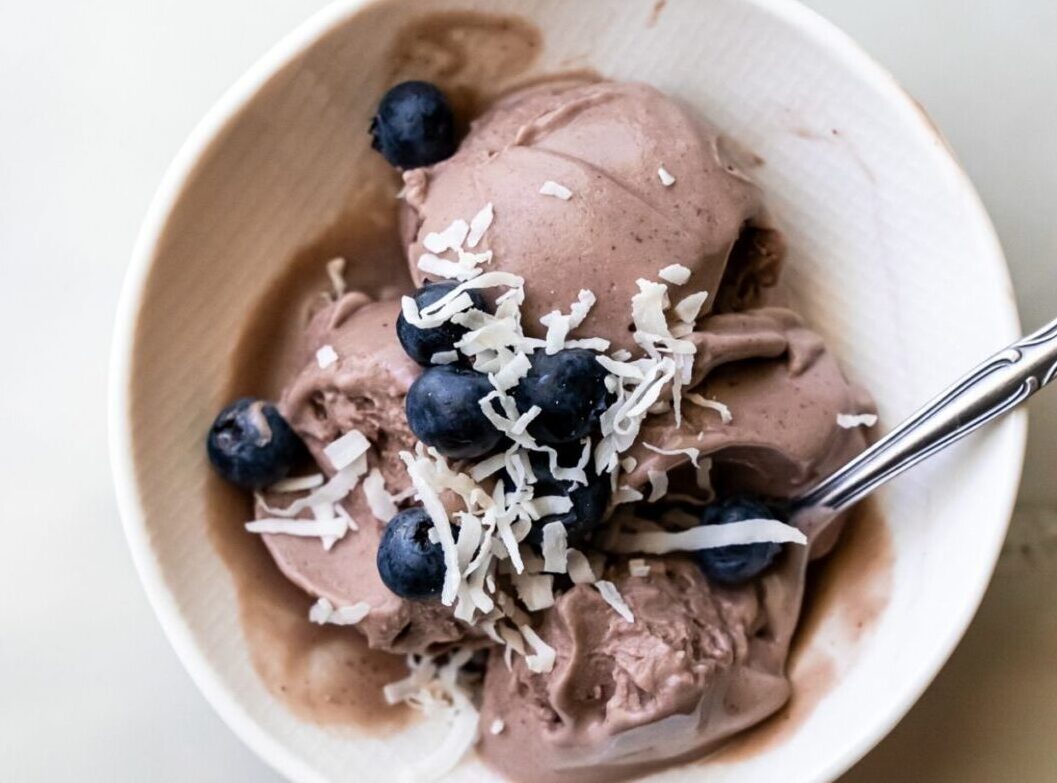 A bowl of chocolate ice cream topped with fresh blueberries and shredded coconut, with a spoon placed inside the bowl, on a light-colored surface; one of our top picks from 25 desserts you'll love.