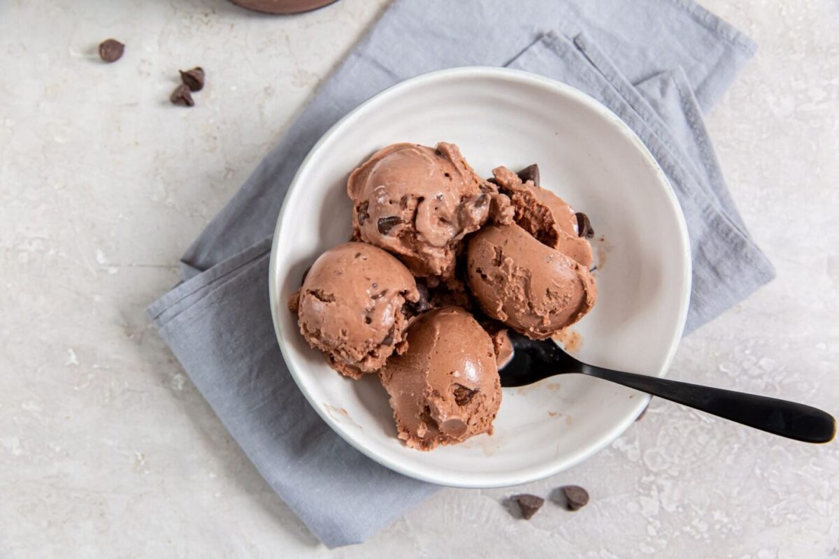 A bowl of chocolate ice cream with chocolate chips, served with a spoon, placed on a folded gray napkin on a light surface.