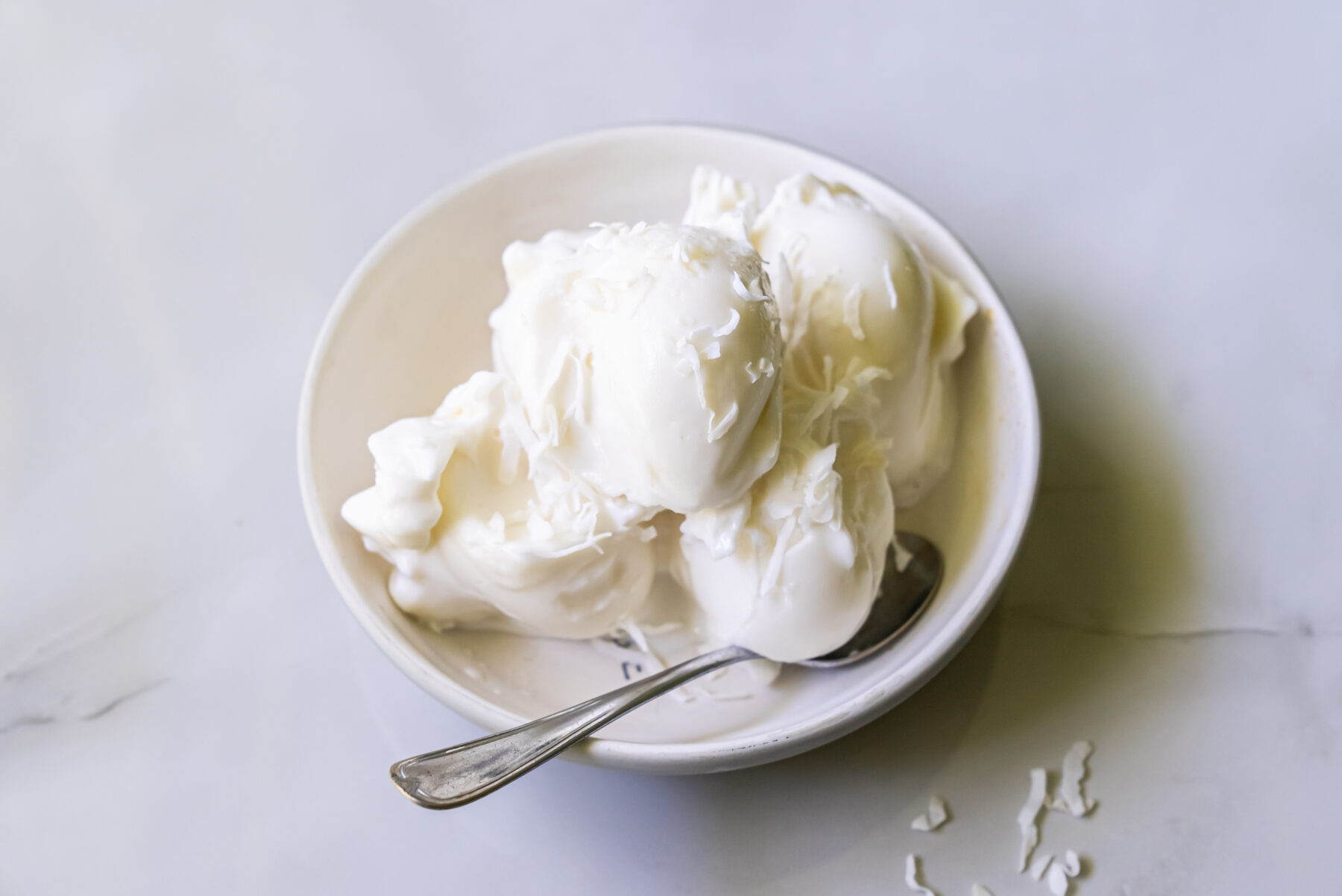 A white bowl filled with scoops of vanilla ice cream, topped with shredded coconut, with a silver spoon placed in the bowl.