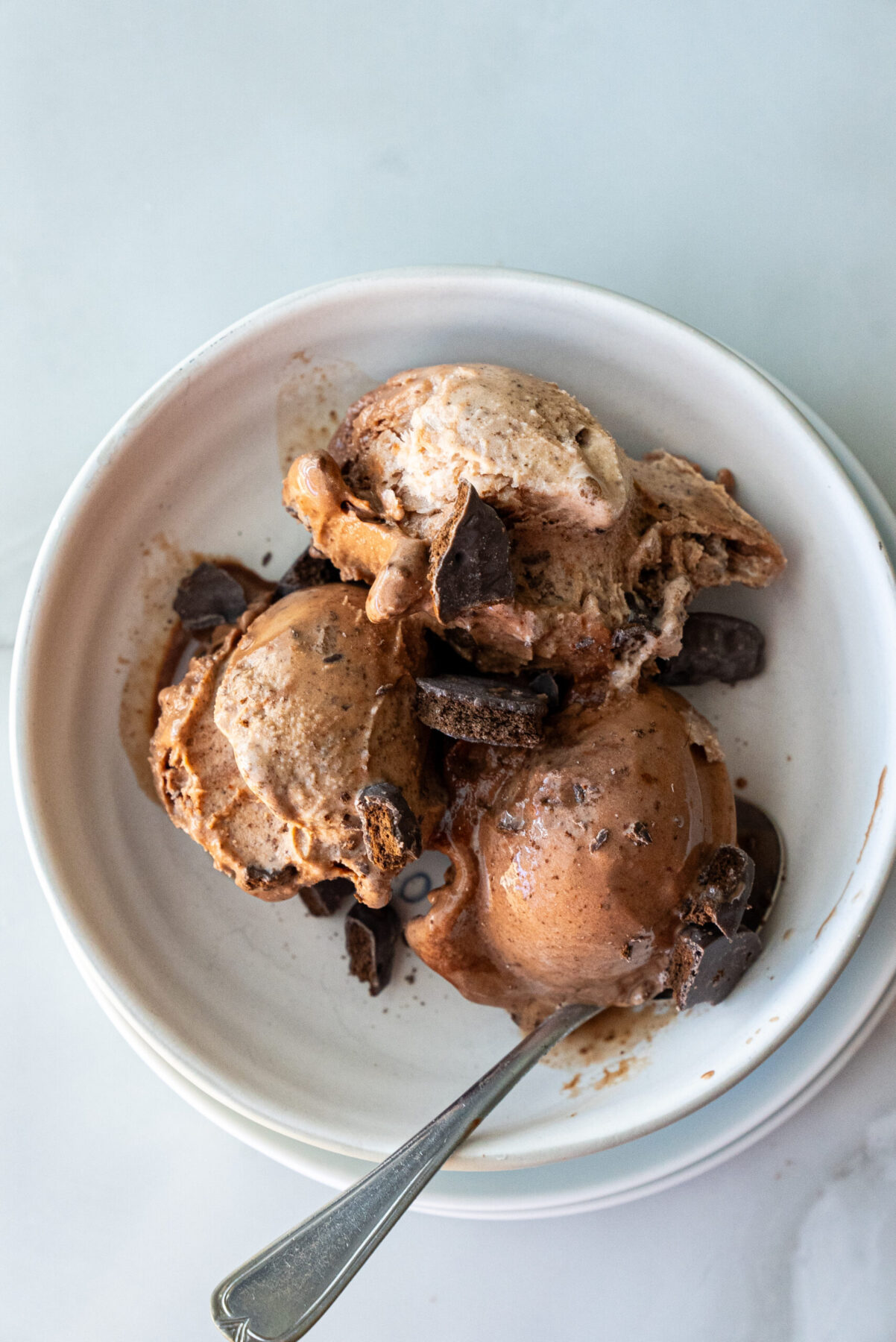 Top-down view of creamy Thin Mint Protein Ice Cream scooped into a white ceramic bowl, studded with crushed chocolate cookies and dark chocolate chunks, with a spoon resting beside.