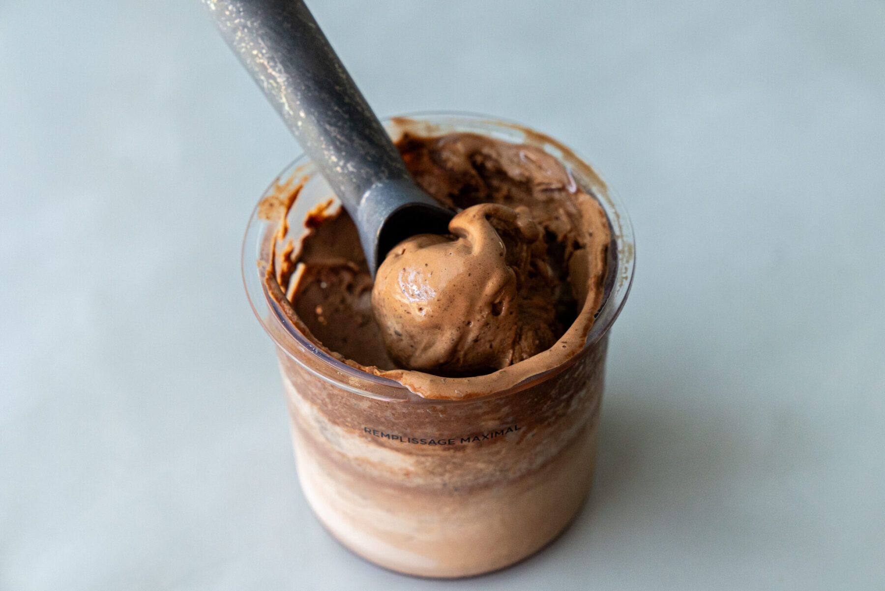 Close-up of a black ice cream scoop lifting a smooth swirl of Thin Mint Protein Ice Cream from a clear Ninja Creami pint, showcasing its dense, fudgy texture speckled with cookie bits.