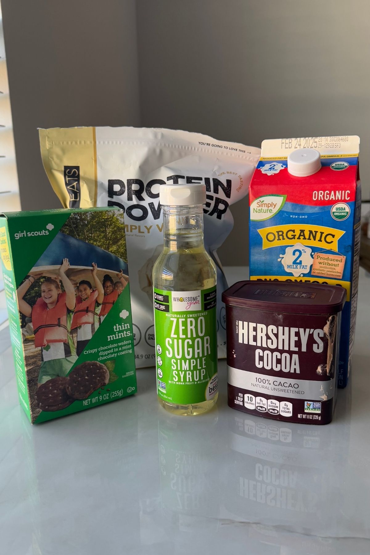 Key ingredients for Thin Mint Protein Ice Cream displayed on marble: Girl Scouts Thin Mints, Hershey’s Cocoa, Simply Nature Organic 2% Milk, Wholesome Zero Sugar Syrup, and Eats Protein Powder — all ready to blend into decadent frozen treat.