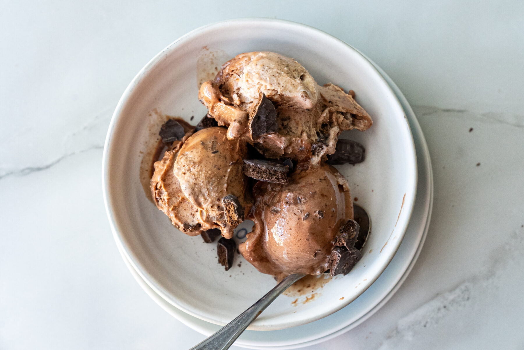 Overhead shot of three generous scoops of Thin Mint Protein Ice Cream in a rustic white bowl atop marble countertop, garnished with crumbled Thin Mints and chocolate shards, spoon inserted.