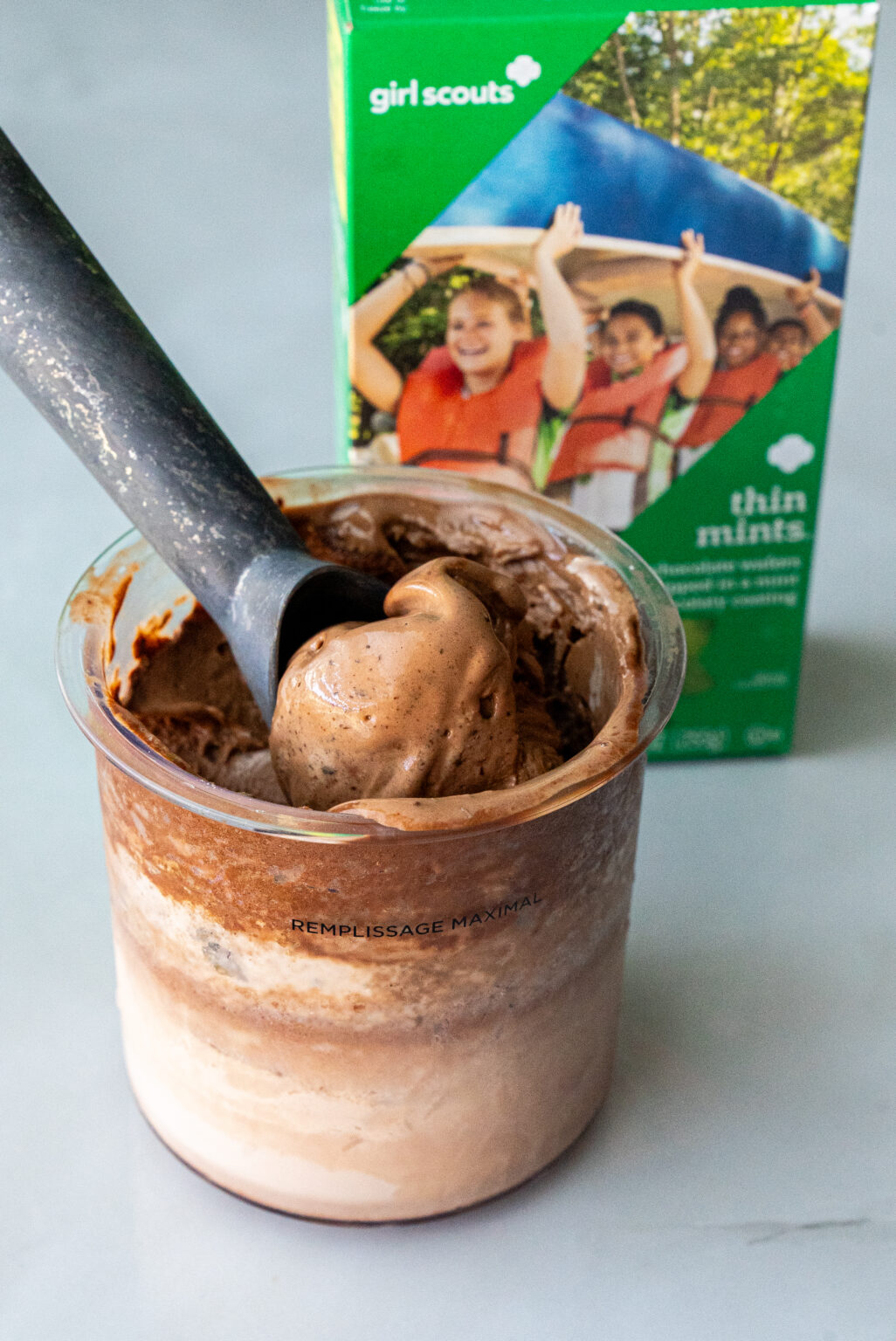 Ninja Creami pint filled with swirled Thin Mint Protein Ice Cream, scoop lifted mid-air, with green Girl Scouts Thin Mints box blurred in background.
