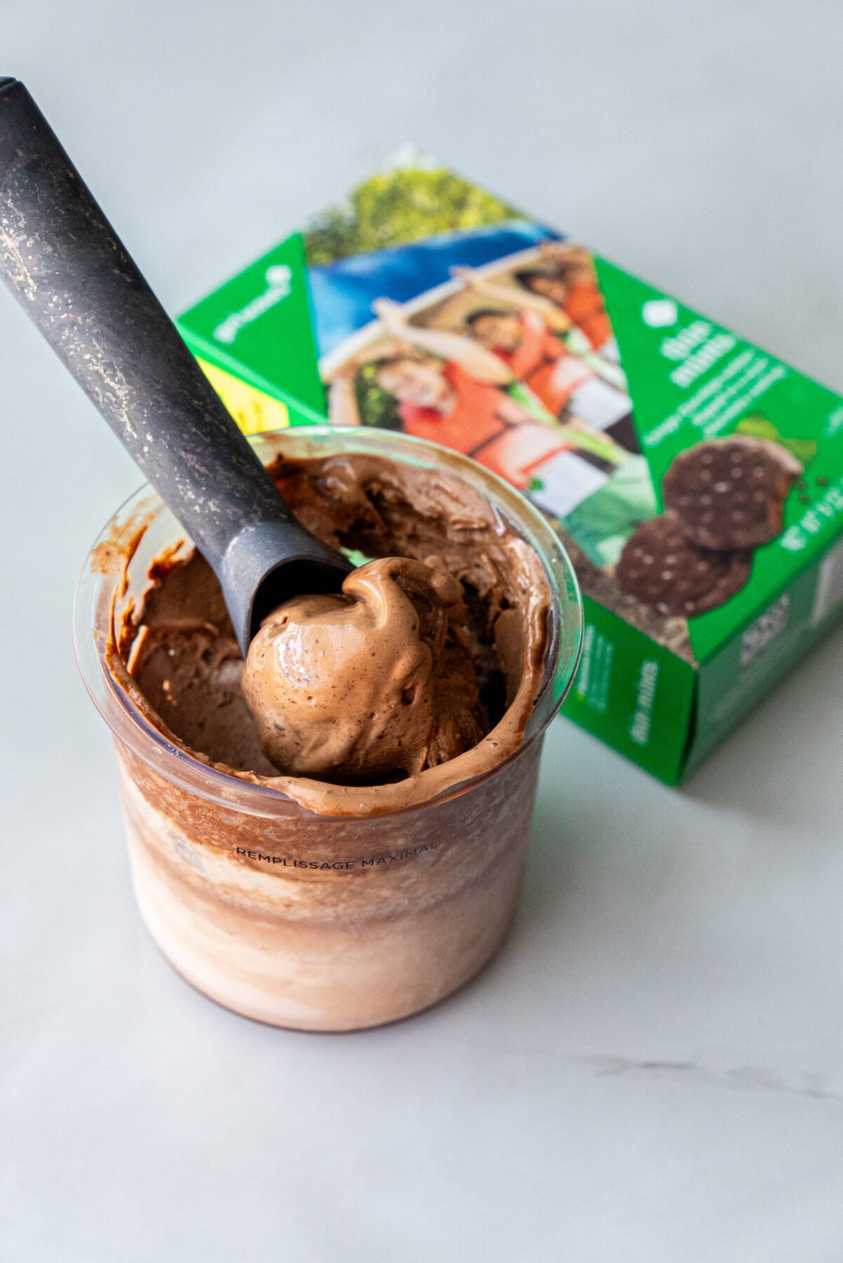 Close-up of creamy Thin Mint Protein Ice Cream in a clear Ninja Creami pint, black scoop lifting a rich swirl, with green Girl Scouts Thin Mints box blurred behind — highlighting mint-chocolate flavor inspiration.