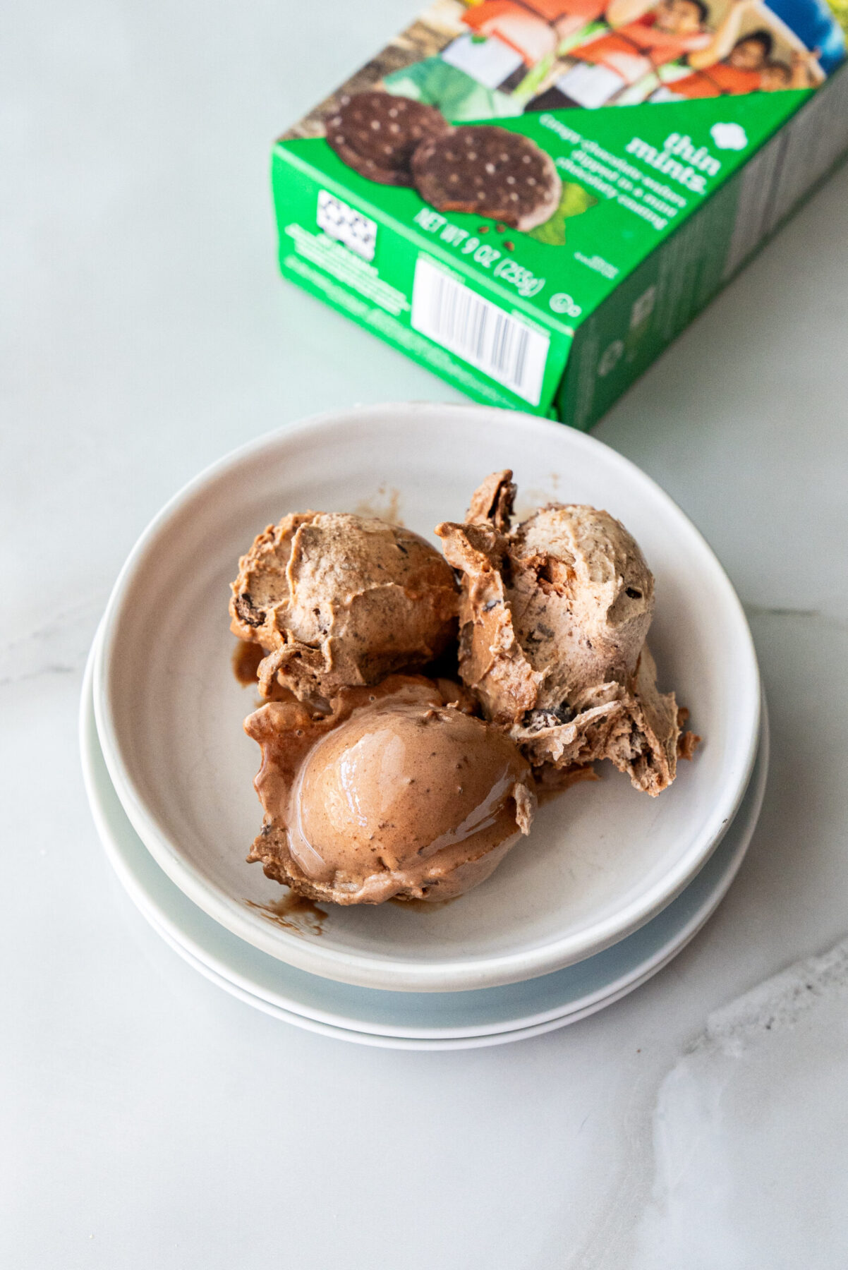 Overhead shot of three melting scoops of Thin Mint Protein Ice Cream in a ceramic bowl on marble, with visible cookie crumbles and glossy sheen; green Thin Mints box angled in background.