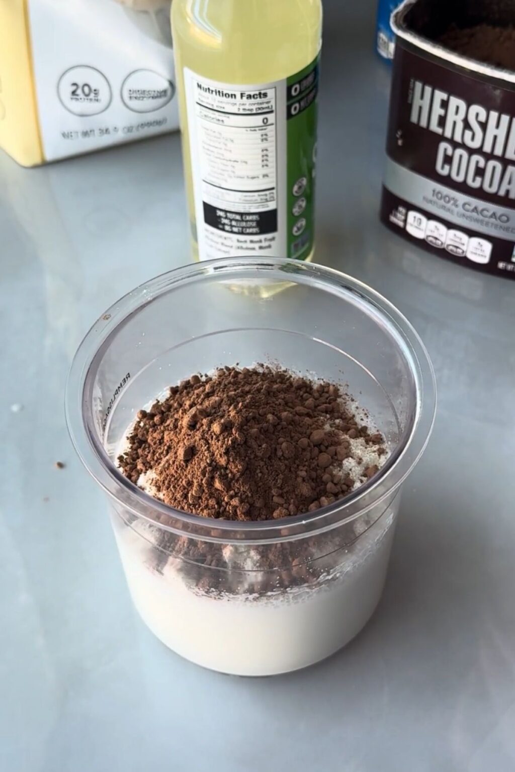 Clear Ninja Creami pint filled with white protein powder topped with Hershey’s cocoa powder, beside a bottle of zero-sugar syrup and protein bag — dry base ready to be mixed into Thin Mint Protein Ice Cream.